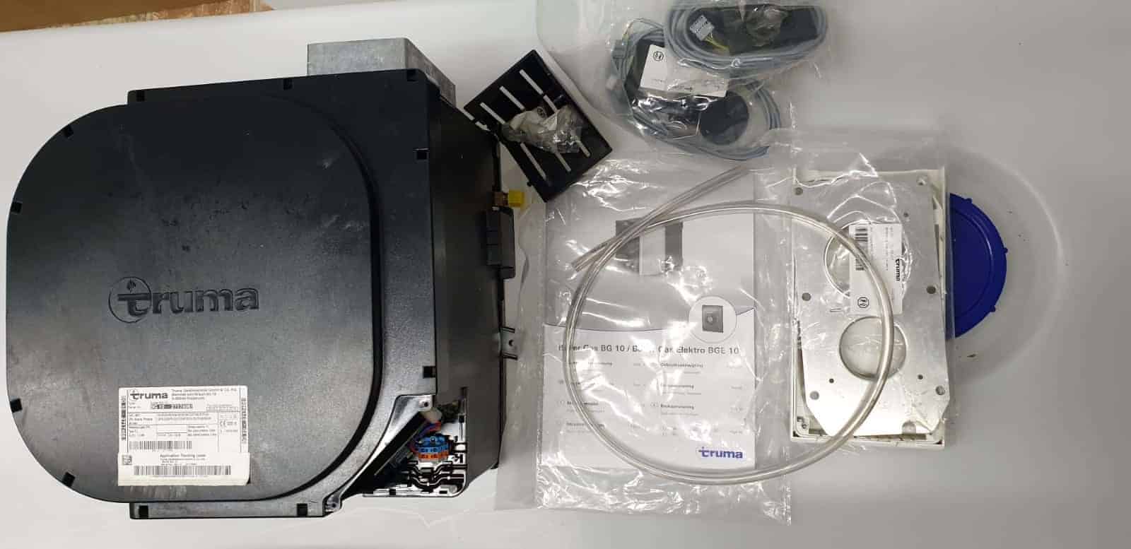 דוד מים לגז וחשמל (TRUMA) עם תווית, חיווט, צינורות, אביזרי התקנה, מדריכים ורכיבי ציוד מחנאות נוספים המוצגים על משטח לבן - אידיאלי לשילוב עם מערכות אנרגיה סולארית, מערכי טכנולוגיה פוטו-וולטאית, פתרונות לייצור חשמל ותשתית אנרגיה מתחדשת.