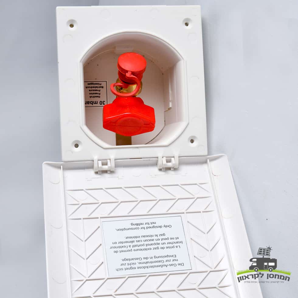 מארז לבן המותקן על קיר עם מכסה פתוח חושף רכיב חיבור חיצוני אדום לגז עבור קרוואן, המשולב בדרך כלל במערכות ייצור אנרגיה סולארית ופוטו-וולטאית שאינן מחוברות לרשת החשמל; תווית הוראות מודפסת נראית על המכסה, ומדגישה חיבור בטוח בתוך תשתית אנרגיה מתחדשת.