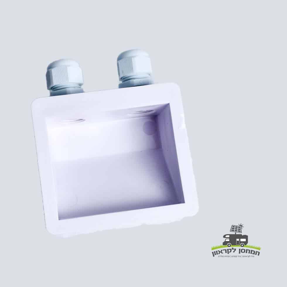 קופסה אנטיגרון אטומה מפלסטיק לבן, עם שני מבלטים לחשמל בחלקה העליון, מיועדת להתקנה בגגות במערכות פוטו-וולטאיות לייצור חשמל סולארי ומתקני אגירה ביתיים באנרגיה מתת. המצולם על רקע אפור בהיר מדגיש את ההתאמה לתשתיות אנרגיה ירוקה. לוגו קטן מופיע בפינה הימנית התחתונה.