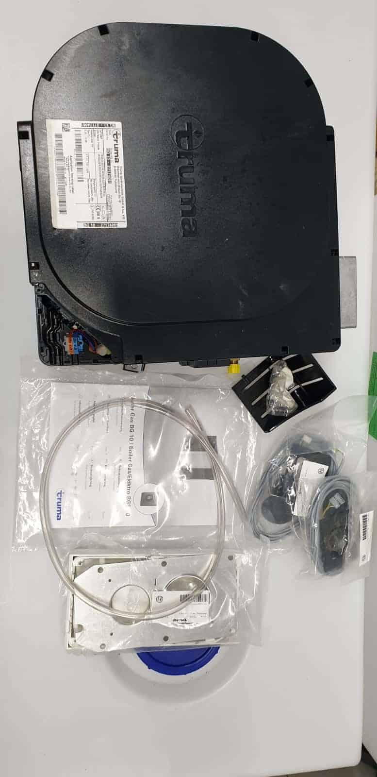 מחמם מים של TRUMA לגז וחשמל עם כבלים, חומרת התקנה ומדריך למשתמש - אידיאלי לציוד מחנאות, תחנות כוח ניידות ושילוב עם פאנלים סולאריים או מערכות פוטו-וולטאיות. אביזרים מוצגים על משטח לבן, תוך שימת דגש על תאימות עם מערכי אנרגיה מתחדשת ותשתית ייצור חשמל בת קיימא.