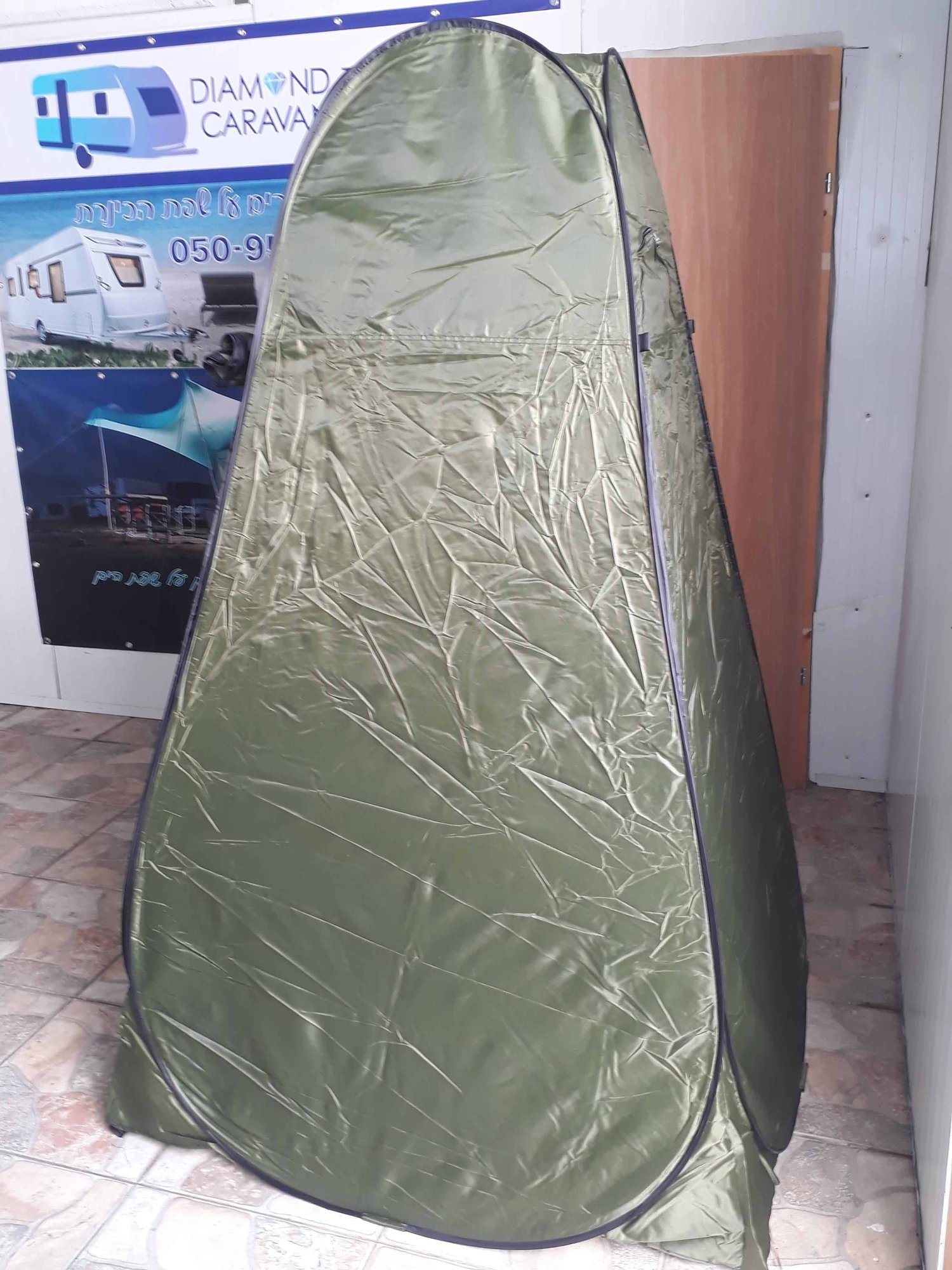 אוהל מקלחת חיצוני מתקפל, אוהל פרטיות מתקפל ירוק, מוצג בתוך הבית על ריצוף אריחים ליד דלת עץ ופרסומת למוצרי קרוואן, המציג את התאמתו לפתרונות אנרגיה סולארית, מערכות פוטו-וולטאיות ניידות, וייצור חשמל מתחדש - החל מחמפינג יעיל מחוץ לרשת ועד אינטגרציה עם תחנות כוח סלולריות חיצוניות בת קיימא.