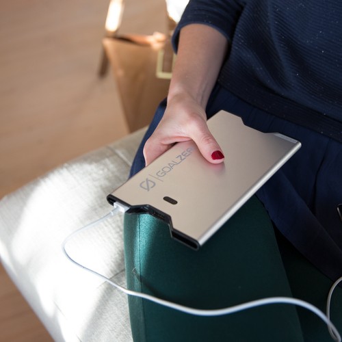 אדם יושב על ספה כשהוא מחזיק סוללת גיבוי מתכתית SHERPA40 (סוללת גיבוי) 40Wh של GOALZERO, מצוידת בכבלים L+M ומחוברת באמצעות כבל טעינה לבן, הממחישת אחסון אנרגיה סולארית ניידת וטכנולוגיית ייצור חשמל פוטו-וולטאית מתחדשת לתשתית אנרגיה בת קיימא.