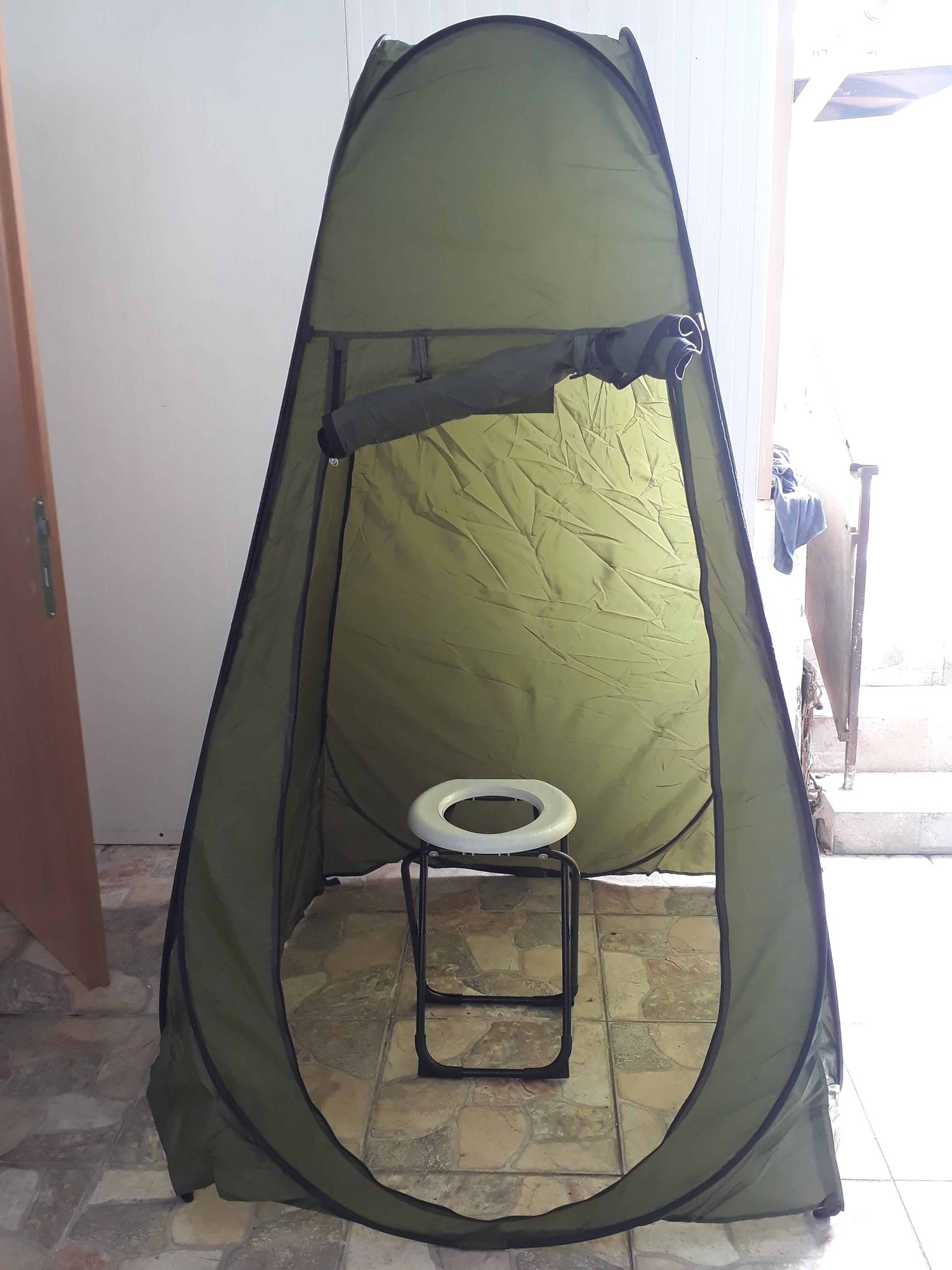 אוהל מקלחת חיצונית מתקפל אידיאלי כציוד קמפינג או לקרוואנים, מוצב ליד דלת פתוחה ומדרגות על פת קרמיקה – מתאים במיוחד לשימוש עם מערכות אנרגיה סולארית, תאורת LED סולארית ותחנות כוח פוטו-וולטאיות ניידות לטובת ייצור חשמל נקי ותשתיות אנרגיה מתחדשת בשטח.