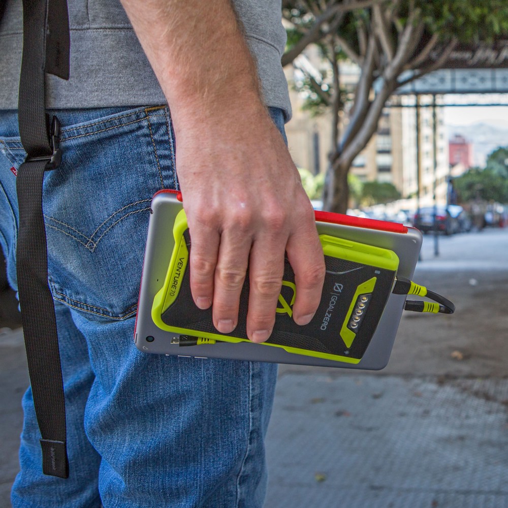 אדם לובש ג'ינס כחול אוחז בטאבלט מוקשח וסוללת גיבוי אדומה מדגם סוללת גיבוי IP67 71Wh VENTURE70, עומד על מדרכה עירונית תוך כדי בדיקת תשתית אנרגיה מתחדשת, כגון תחנת כוח סולארית פוטו-וולטאית או מערכת אגירת אנרגיה סולארית.