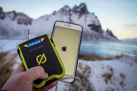 יד אוחזת בסוללת גיבוי סולארית VENTURE30 מדגם IPX6 28Wh המטעינה אייפון בחוץ, מציגה ייצור אנרגיה מתחדשת באמצעות טכנולוגיה פוטו-וולטאית על רקע הרים מושלגים ואגם, תוך הדגשת פתרונות חשמל בני קיימא לסביבות שאינן מחוברות לרשת החשמל.