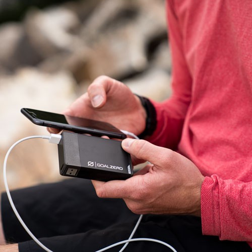 אדם בחולצה אדומה טוען סמארטפון בחוץ באמצעות סוללת גיבוי 36Wh flip36, המציג פתרונות אנרגיה מתחדשת ניידים עם טכנולוגיה פוטו-וולטאית מתקדמת וייצור אנרגיה סולארית לקישוריות בת קיימא מחוץ לרשת החשמל.