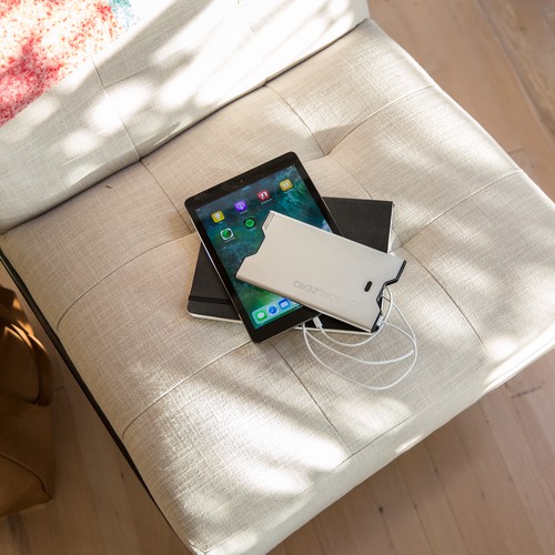 סוללת גיבוי SHERPA40 ניידת בהספק 40Wh עם טכנולוגיה פוטו-וולטאית משולבת ויכולות אנרגיה סולארית, לצד כבלים מחוברים, טאבלט המציג אפליקציות, אוזניות לבנות ומחברת סגורה, כולם מסודרים על כיסא מרופד בצבע בז' - הממחישים אחסון אנרגיה מתחדשת יעיל וייצור חשמל נייד לשימוש טכנולוגי בר-קיימא.