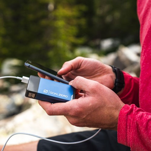 אדם בחולצה אדומה ארוכת שרוולים משתמש בטלפון חכם המופעל על ידי סוללת גיבוי 24Wh כחולה ניידת flip24, המציגה טכנולוגיה פוטו-וולטאית ותשתית אנרגיה מתחדשת לייצור חשמל מחוץ לרשת החשמל בחוץ.