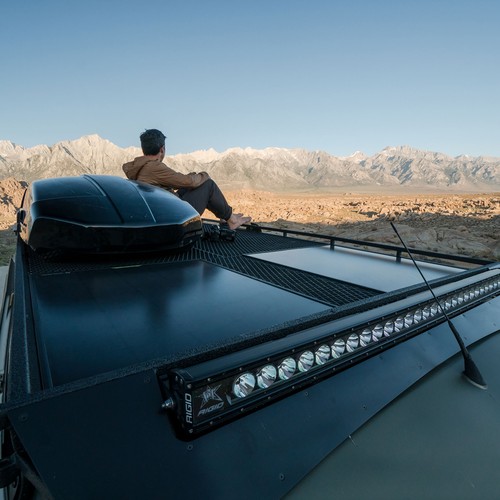 נוסע נח לצד רכבו המצויד בתיבת מטען, ומשקיף על הרים סלעיים תחת שמיים בהירים, ליד פאנל סולארי 12-22V 50W קשיך BOULDER 50. פאנל סולארי פוטו-וולטאי מתקדם זה מנצל אנרגיה מתחדשת מהשמש, מייצר חשמל נקי כדי להפעיל את המסע שלהם ומציג פתרונות חשמל בני קיימא מחוץ לרשת החשמל.