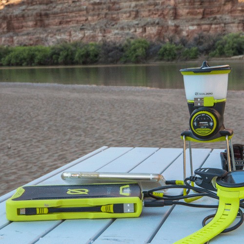 סוללת הגיבוי הניידת VENTURE70 מדגם IP67 71Wh עם גימור צהוב ממוקמת על שולחן לבן ליד גדת נהר חולית, מוקפת בעצים וצוקים סלעיים - אידיאלית לשילוב עם תחנה סולארית כדי לרתום אנרגיה סולארית באמצעות טכנולוגיה פוטו-וולטאית מתקדמת, התומכת בייצור חשמל אמין מחוץ לרשת החשמל ותשתית אנרגיה מתחדשת במהלך הרפתקאות בחיק הטבע.