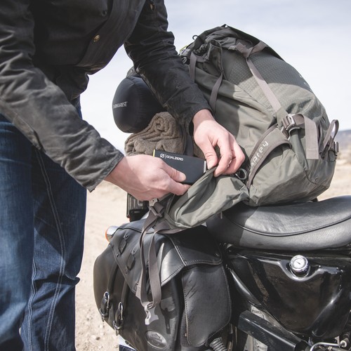 משתמש מניח סוללת גיבוי 15Wh SHERPA15 סוללת קבלים L+M - סוללת גיבוי פוטו-וולטאי מתקדמת המיועדת לאחסון אנרגיה סולארית וייצור חשמל מתחדש - בתוך תרמיל גב אפור של Osprey המותקן על אופנוע בחוץ, ומציג תשתית סולארית ניידת ופתרונות אנרגיה ניידים קיימא.