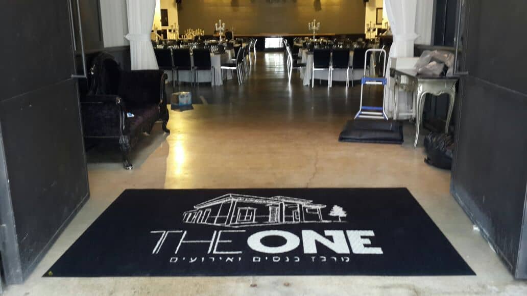 שטיח רצפה שחור עם הכיתוב "THE ONE" ואיור של בניין בכניסה לחדר אוכל גדול ומסודר. מתחת לכותרת הראשית מופיע טקסט בעברית.