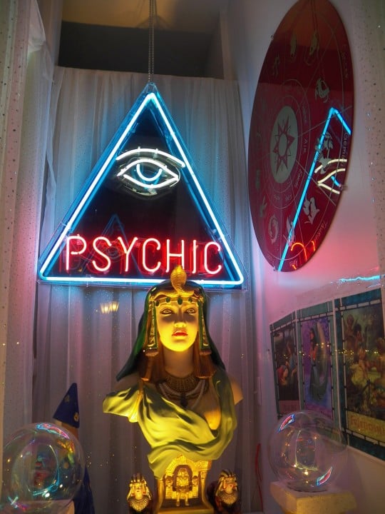 Picture of a glass museum of spiritual tools encased. We see a egyptian head statue of a woman with a red astrology picture on the wall to the right and tarot cards underneath. Crystal quarts balls on both sides and wizard behind.