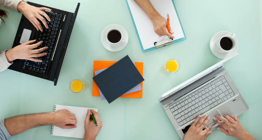Three people work at a table with laptops, notebooks, coffee cups, and orange juice, viewed from above.
