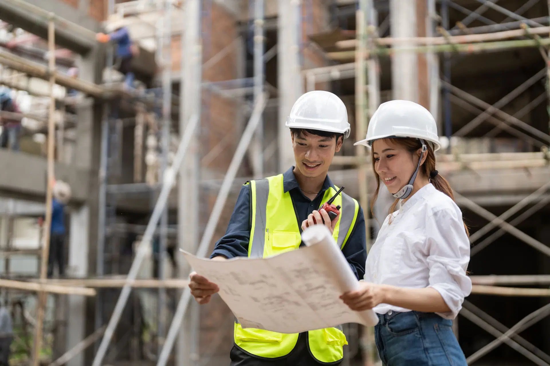 Two construction workers wearing hard hats look at a blueprint and talk on a walkie-talkie at a construction site with scaffolding in the background.