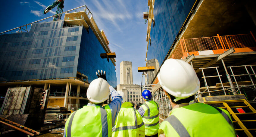 Construction workers in safety vests and helmets observe and point at progress on a construction site with multiple modern buildings under development.