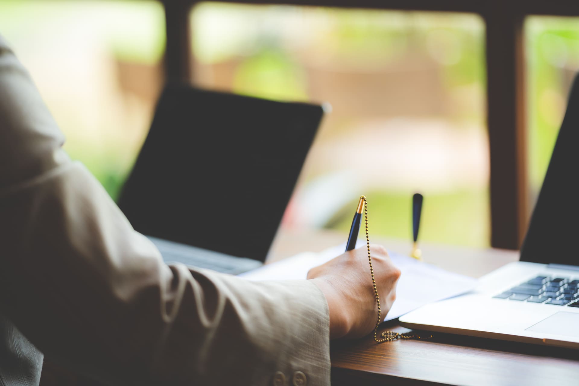 Person in a suit writing on paper at a desk with two laptops, with natural light coming through a window in the background.