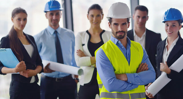 A group of six professionals, some wearing hard hats and holding rolled blueprints, stand together in an office setting.