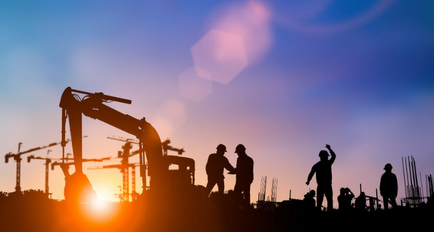 Silhouettes of construction workers and equipment at a building site during sunset, with cranes visible in the background.