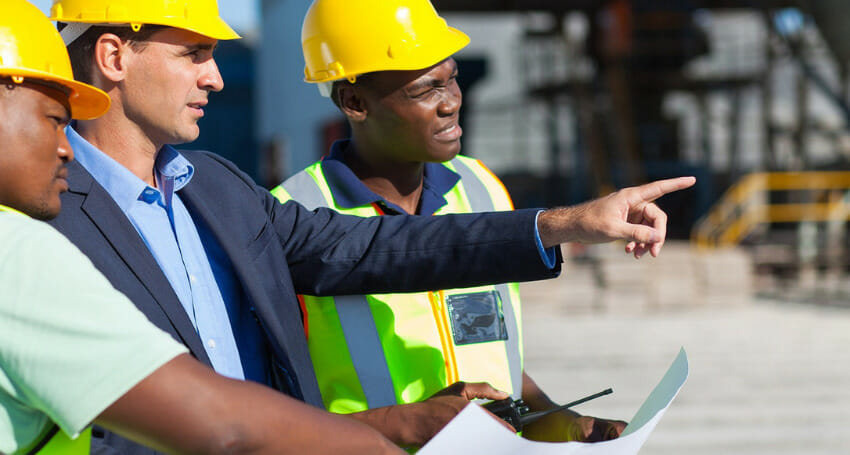 Three men wearing safety vests and hard hats are standing at a construction site, holding documents and discussing as one man points toward something in the distance.