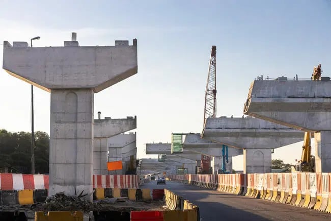 Partially constructed concrete flyover with pillars and beams, roadside barriers, a crane, and a few vehicles on the road below under a clear sky.