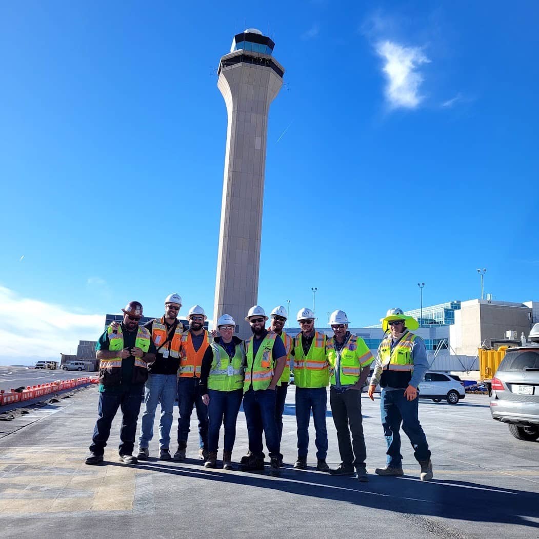 A group of construction workers in safety gear stand together on a tarmac in front of an airport control tower under a clear blue sky.
