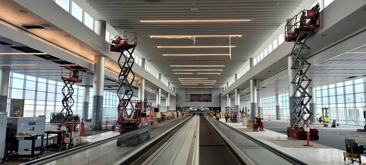 Construction workers on lifts install ceiling fixtures in a spacious, modern airport terminal with large windows and moving walkways.