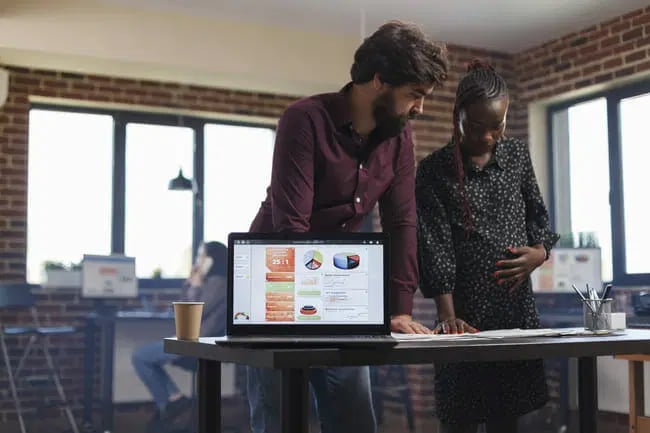 Two people stand at a desk discussing documents, with an open laptop displaying charts and graphics in an office setting.