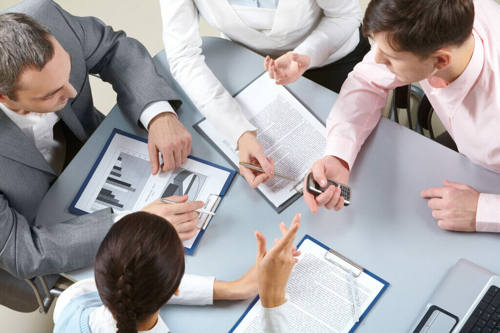 Four people sit at a table discussing documents, charts, and data, with clipboards, papers, pens, and a calculator visible.