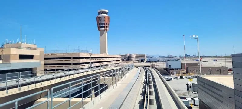 Airport control tower stands above terminal buildings and rail tracks on a clear day, with vehicles and equipment visible on the tarmac.