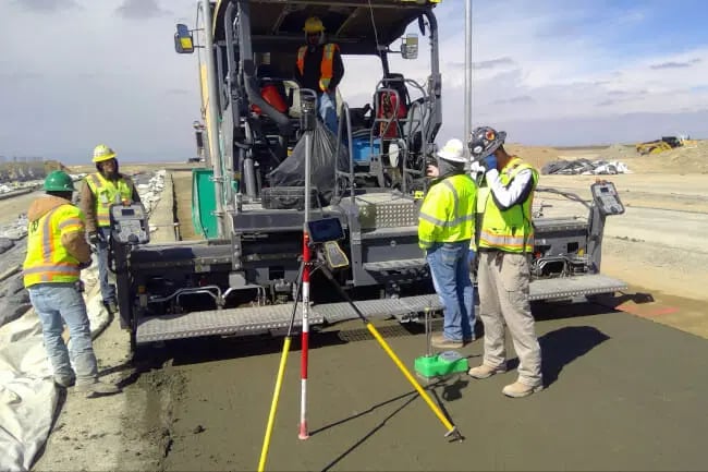 Construction workers in safety gear operate machinery and use surveying equipment on a road construction site under a cloudy sky.