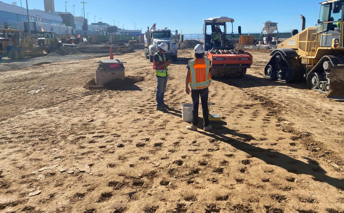 Two construction workers in safety vests and helmets stand on a dirt construction site, surrounded by heavy machinery and vehicles under a clear blue sky.