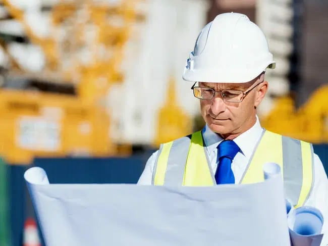A man in a white hard hat and safety vest is looking at blueprints at a construction site.