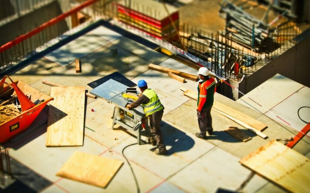 Two construction workers wearing safety gear operate equipment and handle materials on a building site with wood planks and tools scattered around.