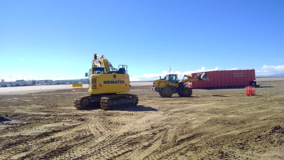 A Komatsu excavator and a front loader operate on a cleared construction site near a row of red shipping containers under a clear blue sky.