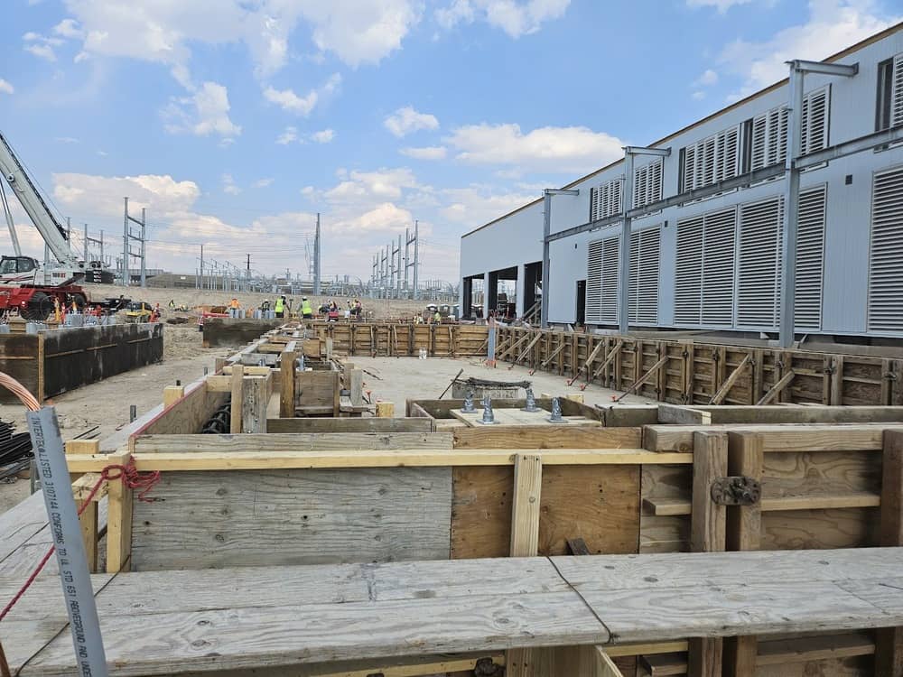 Construction site with wooden framework for concrete, workers in the background, and an industrial building with large vents on the right under a partly cloudy sky.