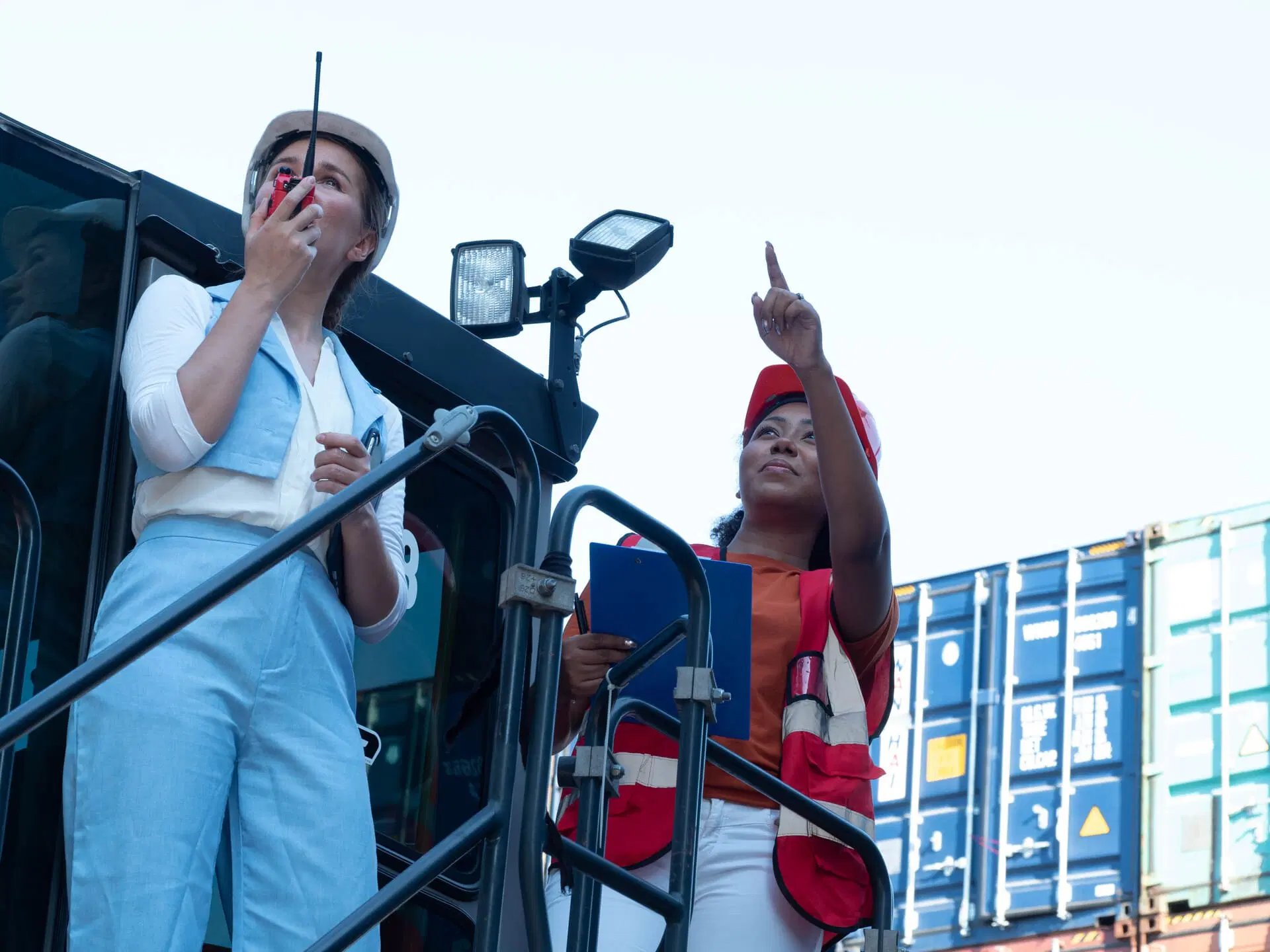 Two women in safety gear stand by shipping containers; one uses a two-way radio, while the other holds a clipboard and points upward.