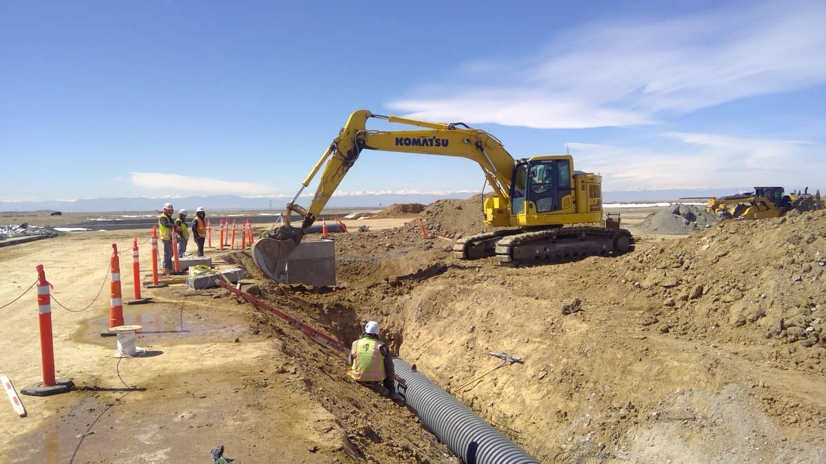A yellow excavator digs near a group of construction workers placing a large black pipe in a trench at a worksite, with orange safety cones lining the area.