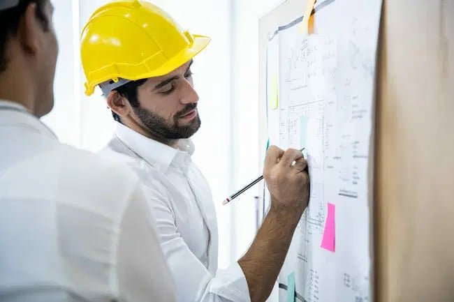 Man wearing a yellow hard hat writes on a large sheet of paper with diagrams and sticky notes, while another person stands nearby.