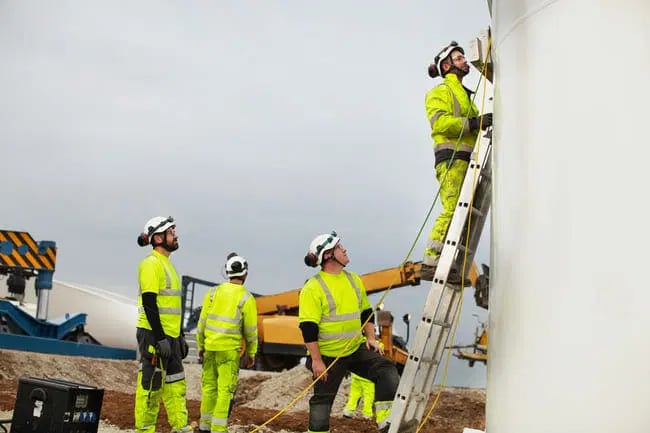 Four construction workers in high-visibility clothing and helmets are working outdoors; one is on a ladder beside a large white structure while three others observe.