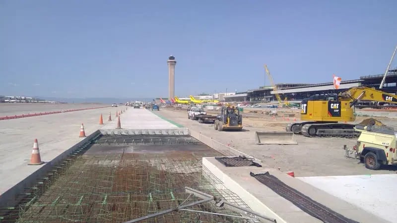 Construction work underway at an airport, with heavy machinery, building materials, traffic cones, and an air traffic control tower visible in the background.