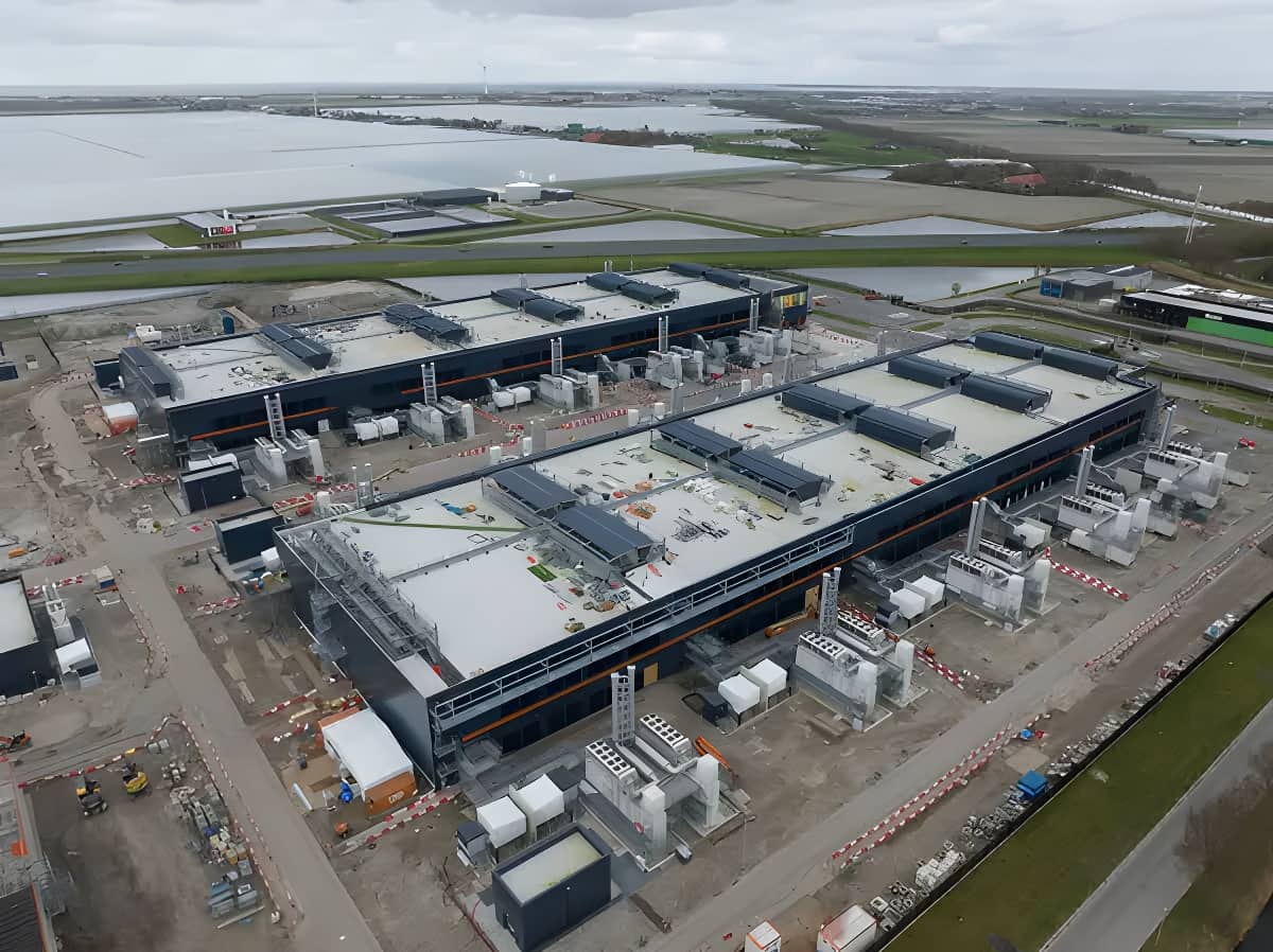 Aerial view of a large industrial facility with multiple rectangular buildings, machinery, and construction activity, surrounded by fields and waterways.