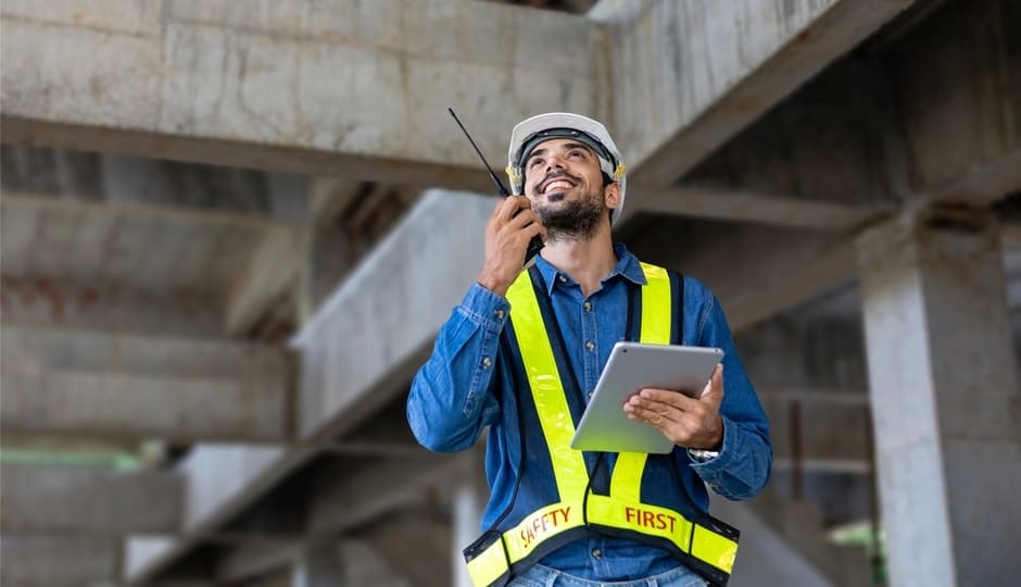 Man in a safety vest and hard hat holds a tablet and talks on a walkie-talkie at a construction site.