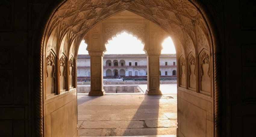 View of a historic courtyard framed by an arched stone doorway with intricate carvings, illuminated by sunlight.