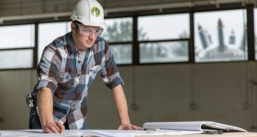 A man wearing a hard hat, safety glasses, and a plaid shirt reviews blueprints on a table inside an industrial building.