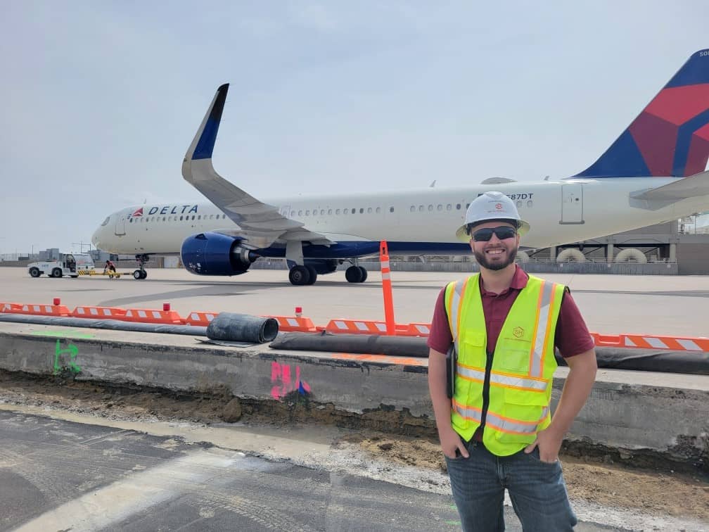 A man wearing a hard hat and safety vest stands in front of an under-construction area near a Delta airplane on an airport tarmac.