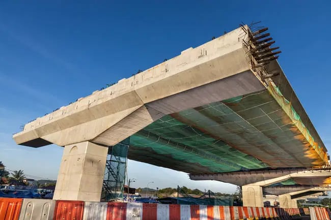 Partially constructed elevated concrete bridge with exposed rebar, supported by pillars, and covered with safety netting beneath; construction barriers are visible in the foreground.