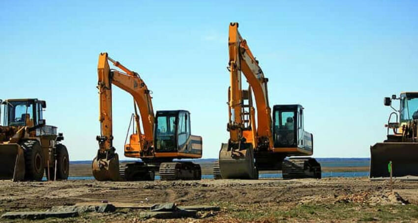 Four large construction excavators and bulldozers are parked on a dirt site near water under a clear blue sky.