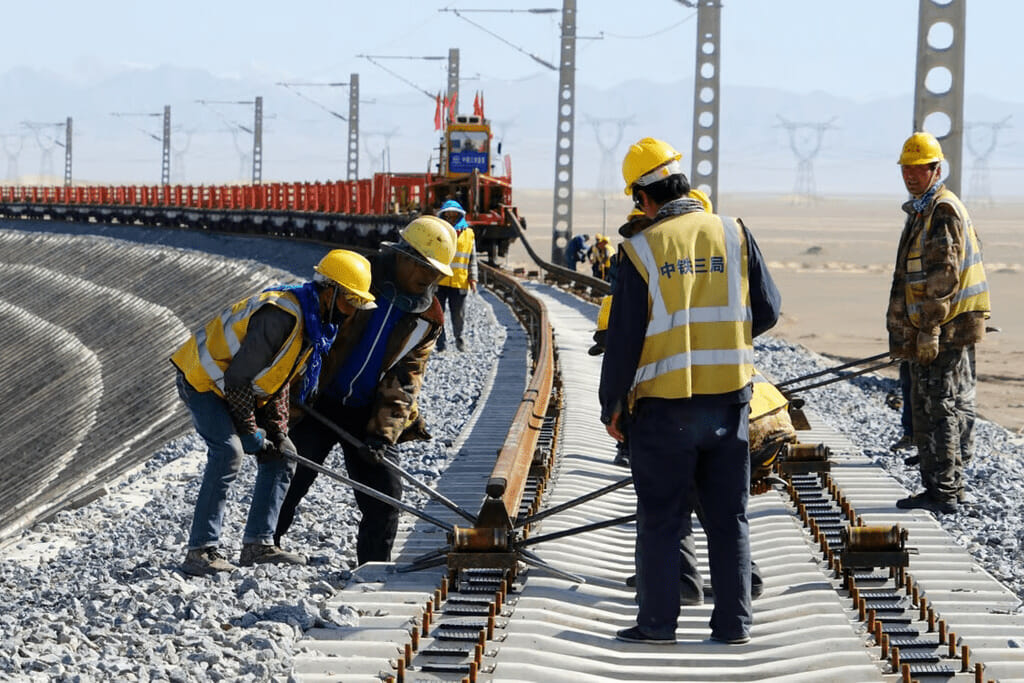 Construction workers in safety vests and helmets are installing railway tracks with tools on a curved section of a new railway line in a remote, open area.