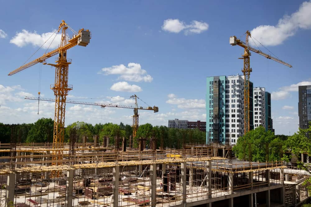 Construction site with multiple cranes and partially built structures in front of modern residential buildings under a blue sky with clouds.